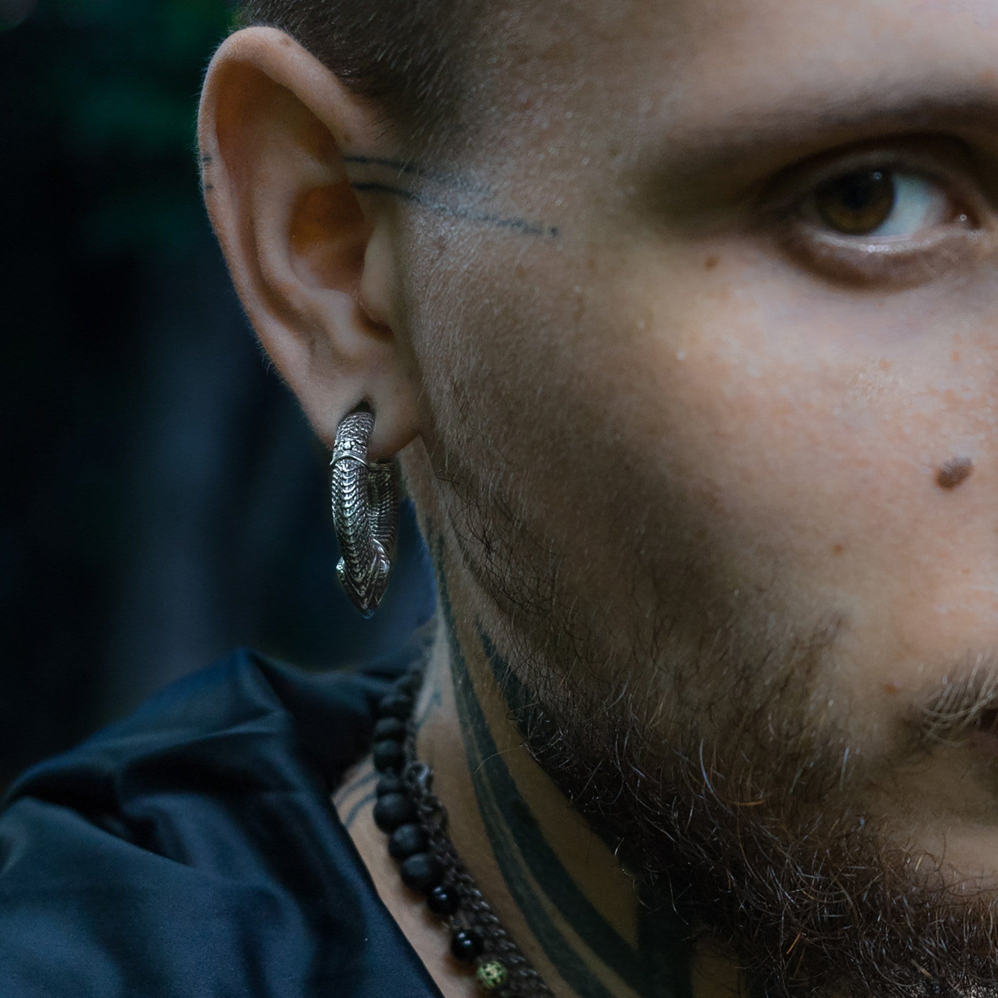Close-up of the right side of a man's face, showcasing the MARVDASHT Small Biomechanical Clicker Ear Weights in 925 Silver (2 gauge) by Kaya Movement, beautifully nestled in his ear and giving major sci-fi vibes. He sports tattoos near his ear and on his neck, paired with a beaded necklace around his neck. The dark, out-of-focus background adds to the futuristic feel.
